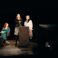 Na ciemnej scenie dwie kobiety stoją przy mównicy, jedna z nich mówi do mikrofonu. Po lewej dwie kobiety siedzą przy czarnym kubiku.