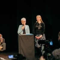 Widok ciemnej sceny. Przy podeście stoją dwie kobiety, jedna z mikrofonem. Po lewej siedzi kolejna kobieta, po prawej niewyraźny kamerzysta.