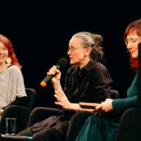 Trzy kobiety sfotografowane z boku. Jedna z nich, siedząca w środku trzyma mikrofon i coś mówi.
