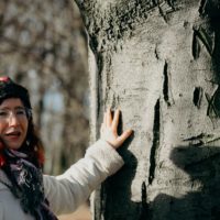 Kobieta w okularach i zimowej czapce stoi przy grubym pniu drzewa. Mówi coś, a wyciągniętą dłonią dotyka pnia.
