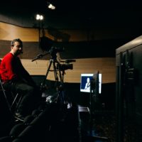 Mężczyzna w czerwonym swetrze siedzi na wysokim krzesle przed kamerą. Przed nim ekran, na którym widać widok z kamery.