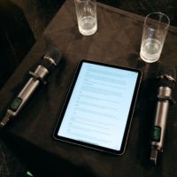 Na czarnym stoliku tablet z wyświetlonym tekstem, dwie szklanki i dwa mikrofony.