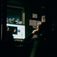 Mężczyzna podparty na łokciu przy biurku i patrzy w ekran, na którym rozmawiają ze sobą dwie osoby. Kadr jest bardzo ciemny, oświetlony punktowo.