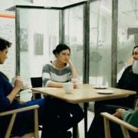 Cztery kobiety siedzą przy drewnianym stoliku. Częśc pije kawę, jedna z nich je ciastko z talerzyka. W tle przeszklenie, za nim fragment wystawy.