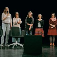 Pięć kobiet stoi na scenie. Jedna z nich, skrajnie po lewej, trzyma mikrofon i coś mówi. Przed nimi stoja puste fotele i nieduże stoliki.