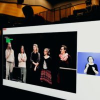 Na ekranie: pięć kobiet stoi na scenie. Jedna z nich, skrajnie po lewej, trzyma mikrofon i coś mówi. W niedużym okienku tłumaczka migowa.