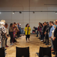 Kilkadziesiąt kobiet stoi w dwóch rzędach naprzeciwko siebie, patrzą na stojącą na środku kobietę w kręconych włosach i żółtej bluzie. Jedna z kobiet na samym końcu prawego rzędu przesuwa krzesło.